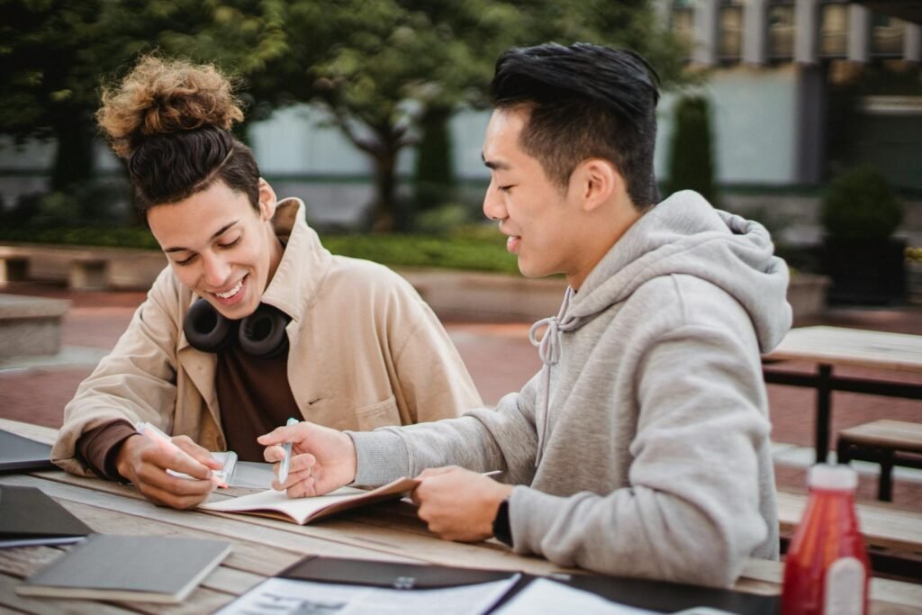 Cheerful multiracial male students wearing casual outfit working on college project together while sitting at wooden table in campus park